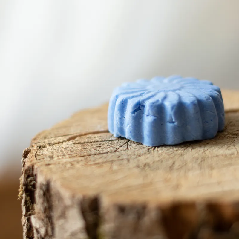 Ein blaues, blütenförmiges festes Shampoo auf einem rustikalen Holzstumpf. Das minimalistische Setting mit natürlichem Licht unterstreicht die Nachhaltigkeit und Reinheit des Produkts, perfekt für eine bewusste Haarpflege.